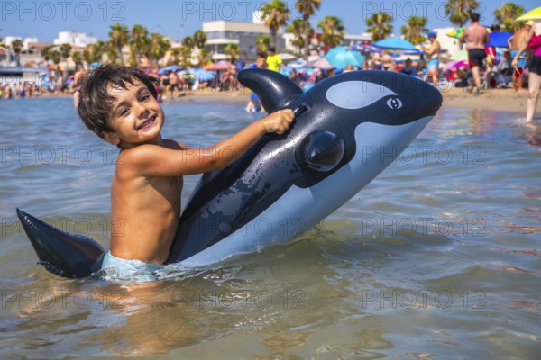 Smiling child having fun riding an inflatable orca toy in the shallow sea water by the beach on a sunny summer day