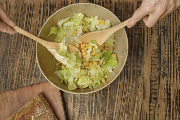Hands are stirring a bowl of fresh salad featuring crisp lettuce and sweet corn. Nearby, a rustic wooden board holds a piece of crusty bread, suggesting a delightful meal experience