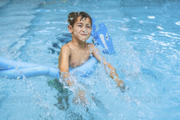 Smiling young boy splashing in a swimming pool, using a blue pool noodle. Joyful summer activity and childhood fun in the water