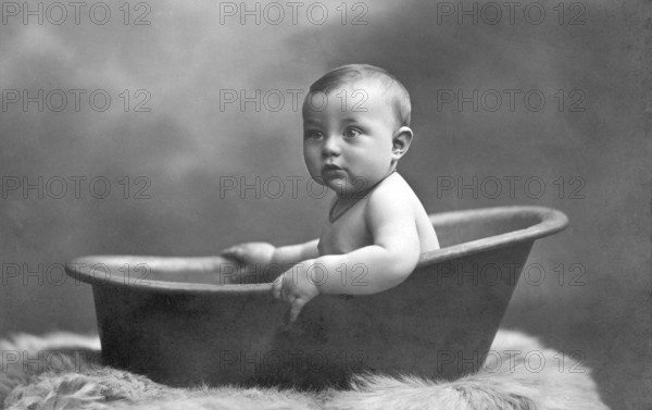 Baby in bathtub, 1930s, A baby sits in an old bathtub holding on to a fur rug, Historical photo