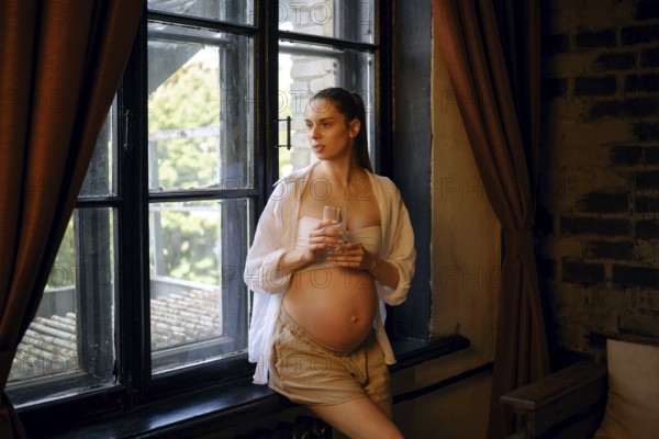 A pregnant woman stands by a large window in her home, holding a glass of water. She gazes outside, reflecting on her journey. Soft light filters through the curtains, creating a serene atmosphere