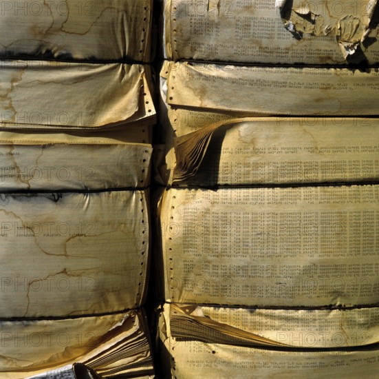 A large stack of compressed paper products is arranged in a recycling area. The paper displays wear and tear, indicating its previous use. This setup highlights recycling efforts