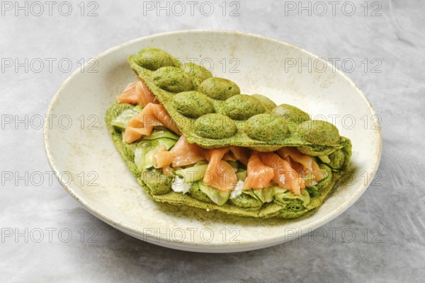 A vibrant green matcha waffle sandwich is filled with fresh salmon, crisp vegetables, and creamy spread. This dish is presented on a neutral plate, highlighting its colorful ingredients and textures