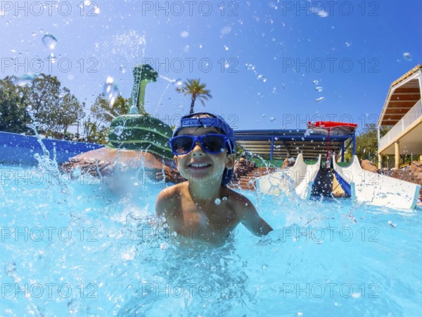 Young boy wearing blue sunglasses, joyfully splashing in a swimming pool at a vibrant water park, embracing summer fun and adventure