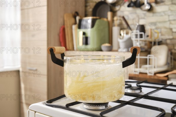 A glass pot filled with boiling water and pasta on a stovetop in a contemporary kitchen. The cozy space features wooden cabinets and various kitchen tools on display