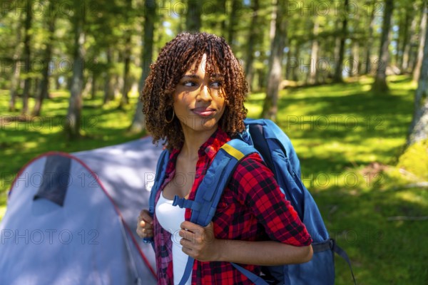 Young woman with a backpack enjoying the tranquility of a forest, discovering nature while camping near a cozy tent on her adventure