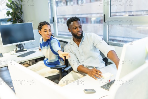 Two colleagues are working together in a modern office, discussing a project on a computer, pointing at the screen and sharing their expertise