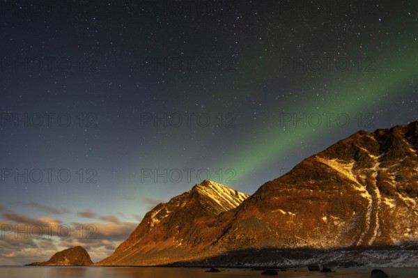 Aurora borealis in green colors over mountain range and coastline at night, Lofoten, Norway