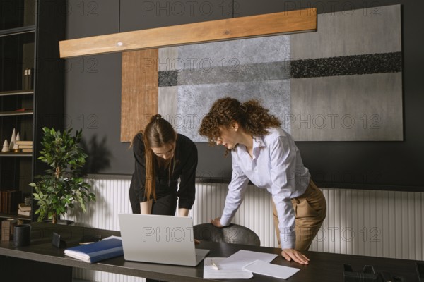 Two women discuss a project at a contemporary office desk, engaging with a laptop and scattered documents. The environment features sleek decor and plants, promoting a productive atmosphere