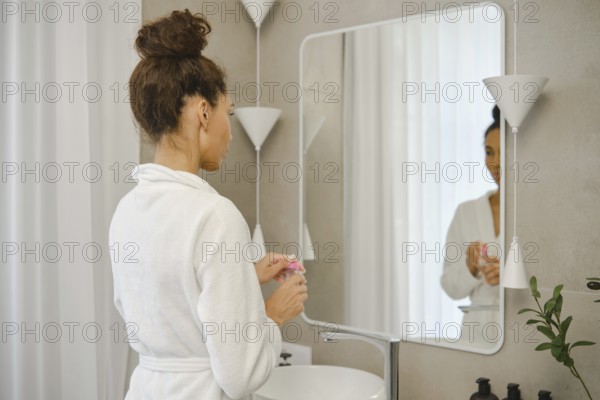 A woman wearing a white robe stands in front of a large mirror in a stylish bathroom. She opens a jar of micellar water and looks at her reflection