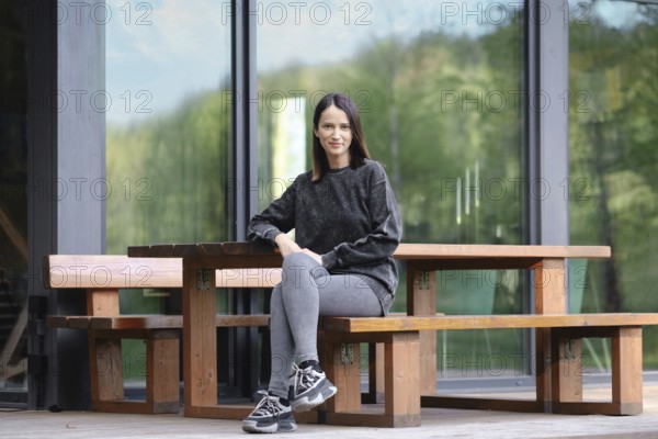 A young woman sits comfortably at a large wooden table placed outdoors. She wears a casual outfit consisting of a sweater and leggings, enjoying her time in a serene environment surrounded by trees
