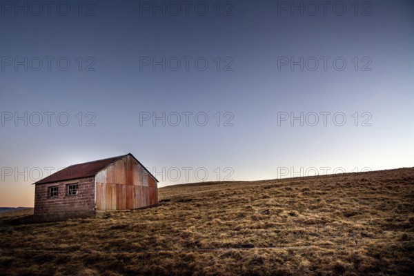An isolated wooden hut sits on a grassy slope in the Regional Nature Park of Volcans d'Auvergne, surrounded by a serene landscape at dusk. The sky transitions from daylight to twilight, Puy de Dome, Auvergne Rhone Alpes, France