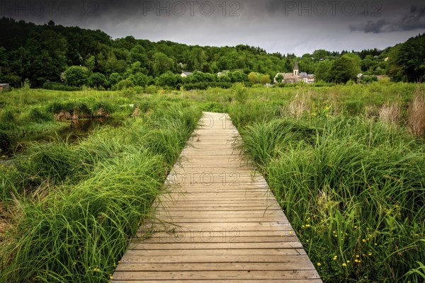 A wooden path stretches through vibrant green grass in the countryside of Auvergne, France. Distant hills and small village details are visible under a cloudy sky, inviting exploration