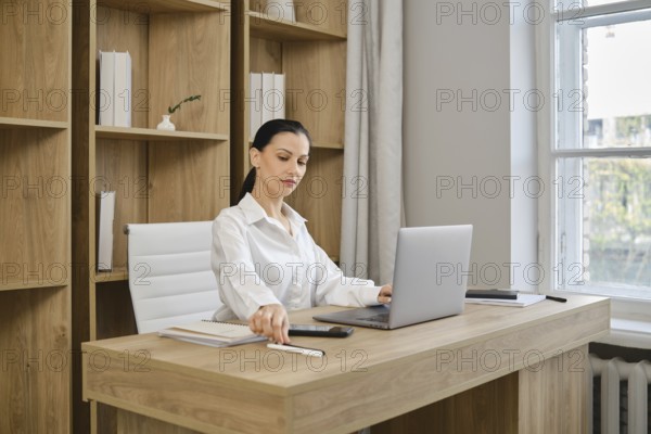 A businesswoman working at a desk with a laptop reached for a pen to make a note