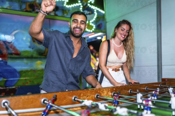 Young man and woman enjoying a foosball game at a funfair, celebrating a goal with excitement and joy
