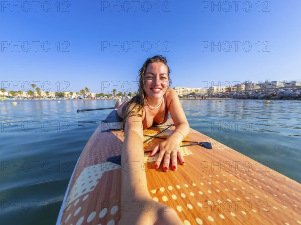 Smiling woman lying on a paddle surfboard, capturing a selfie with a stunning beach and clear blue sky in the background