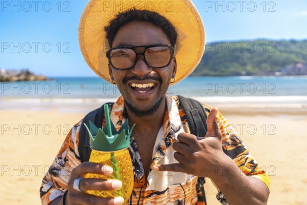 Young man enjoying summer vacation on a tropical beach, holding a pineapple shaped cocktail and gesturing the shaka sign