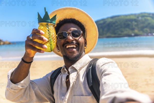 Young man wearing sunglasses and straw hat holding pineapple shaped cocktail while taking selfie on beach