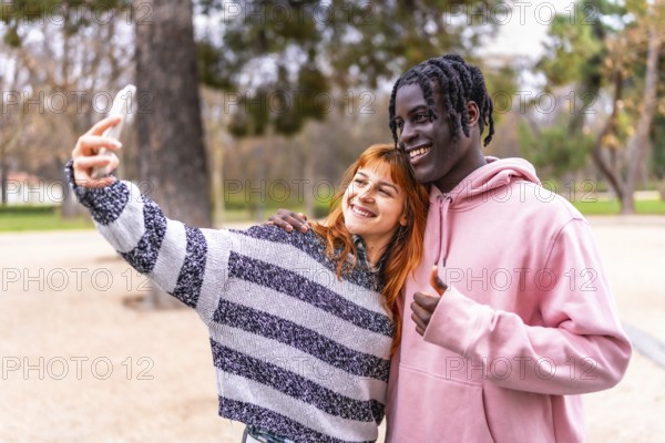 Happy multi ethnic couple enjoying a sunny day in the park, taking a cheerful selfie while flashing thumbs up signs and smiling brightly