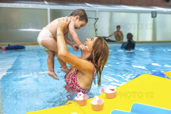 Mother playfully lifting her smiling baby out of the water in an indoor swimming pool during a baby swimming lesson