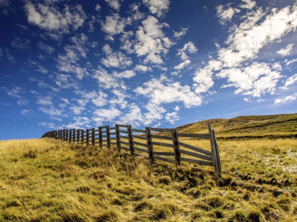 A wooden barrier runs across a grassy area on Sancy mountain, part of the Auvergne Volcanoes Natural Park. Bright clouds drift in a clear blue sky, enhancing the serene landscape. Puy de Dome. Auvergne Rhone Alpes. France