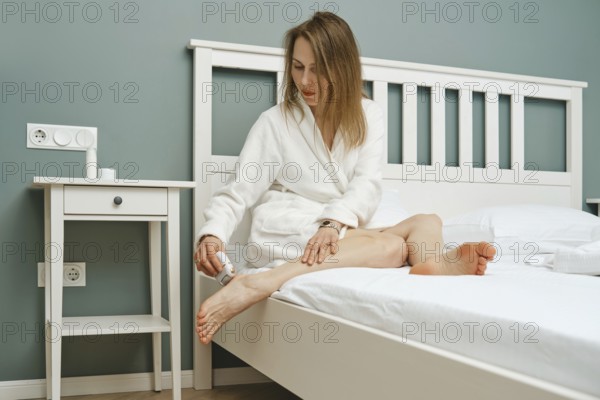 A woman in a white robe sits on the edge of a bed, using electric exfoliating machine on her bare heel. The calming turquoise walls and simple furniture create a relaxing atmosphere