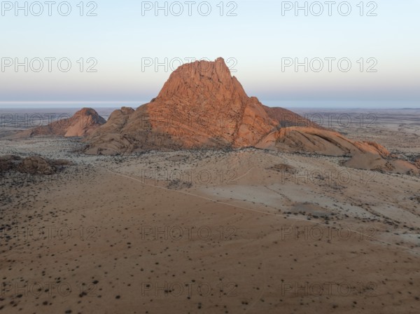 The isolated Spitzkoppe mountain (1728 m) majestically rises above the surrounding desert plains. In the distance the coast and the Atlantic Ocean. At dawn. Aerial view. Drone shot. Damaraland, Namibia