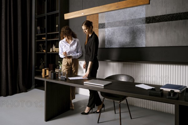 Two women engage in a thoughtful discussion in a sleek office setting. One takes notes on a clipboard while the other points to a document on the table, showcasing teamwork and professionalism