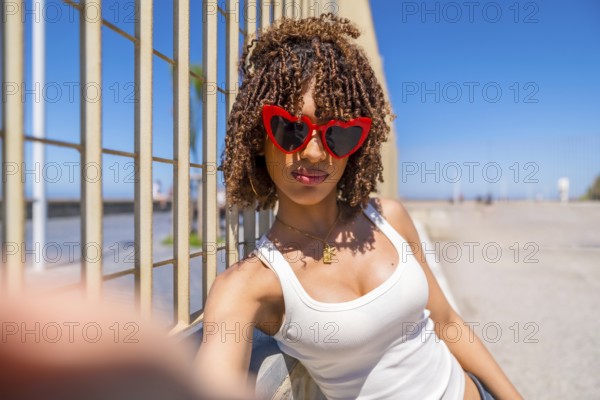 Fashionable young woman with curly hair taking a selfie by the beach, wearing trendy heart shaped sunglasses and a white top