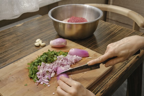 A person chops purple onions and fresh herbs on a wooden cutting board. A bowl of ground meat sits in the background, ready for cooking