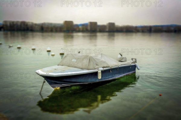 A solitary boat sits anchored in still waters, surrounded by gentle mist. The shoreline is faintly visible in the background, creating a serene atmosphere at dawn, Annecy, Haute Savoie, Auvergne Rhone Alpes, France