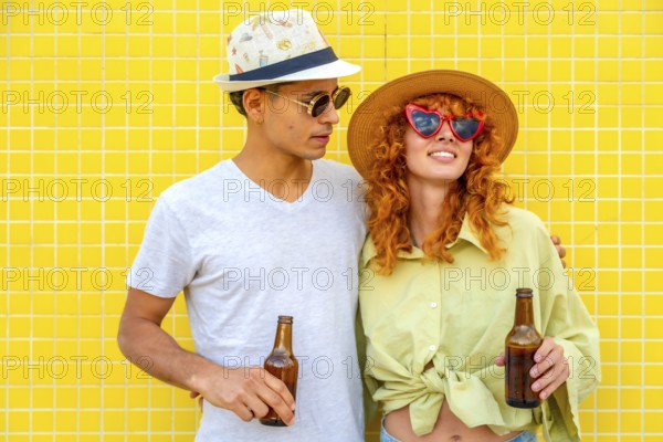 Young couple wearing hats and sunglasses, holding beer bottles and smiling joyfully against a vibrant yellow tiled wall, radiating happiness