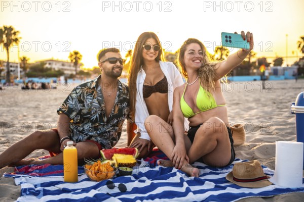Three friends are enjoying a beach picnic at sunset, taking a selfie with fresh fruit and snacks