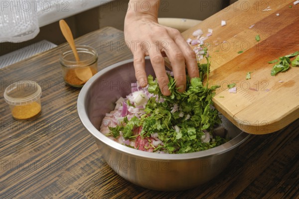 A hand adds freshly chopped cilantro and onions into a large metal bowl. The cozy kitchen countertop is adorned with spices and utensils, suggesting a culinary adventure in progress