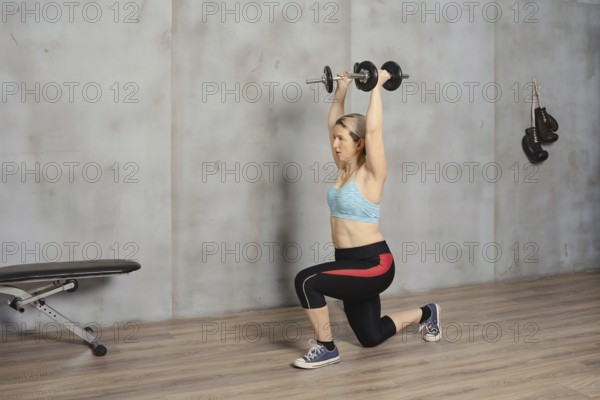 A woman is engaged in a strength training workout, lifting dumbbells overhead while in a lunge position. She is inside a gym, wearing athletic clothing, focusing on her fitness routine