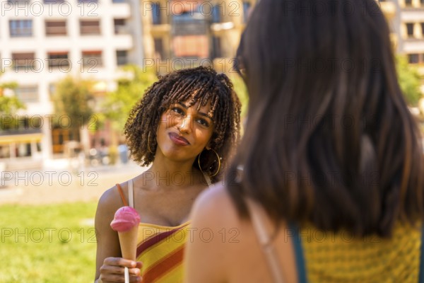 Young woman enjoying a strawberry ice cream cone while talking to her friend in a city park on a summer day