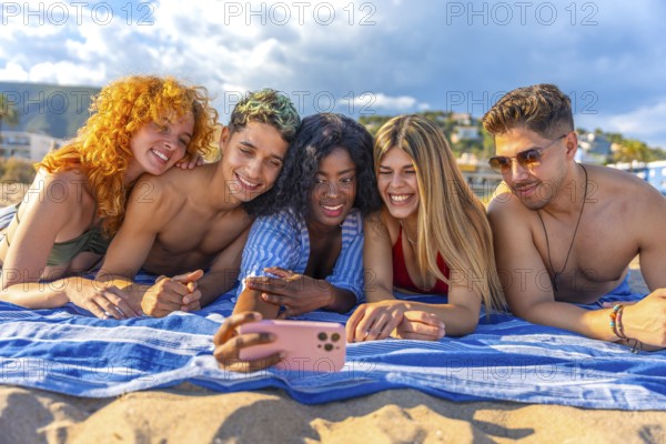 Five cheerful young friends are lying on a beach towel, taking a selfie with a smartphone and enjoying their summer vacation