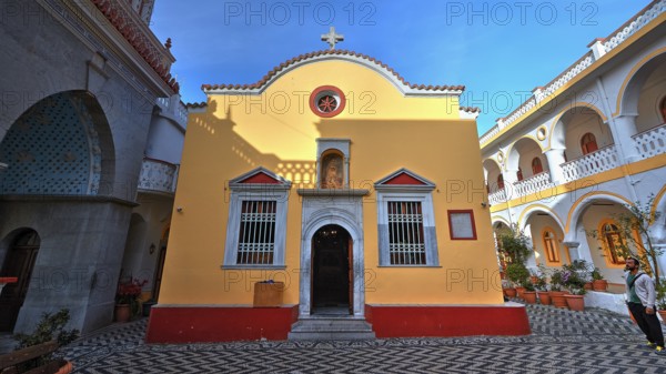 Yellow church with courtyard, cross and baroque elements, surrounded by plants, Holy Week, Bay of Panormitis, Panormitis Monastery, Symi, Dodecanese, Greek Islands, Greece