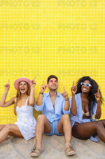 Group of cheerful multi ethnic friends pointing up while sitting on the sand against a vibrant yellow tiled wall, enjoying their summer vacation together