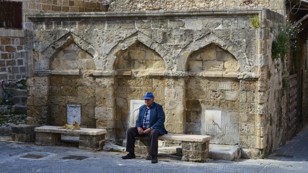 Man with blue hat sitting in front of a ruin-like facade with benches, fountain, Rhodes Old Town, Rhodes, Dodecanese, Greek Islands, Greece