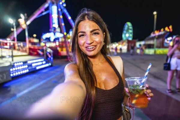 Happy young woman enjoying a vibrant night at an amusement park, taking a selfie while holding a colorful cocktail and smiling brightly