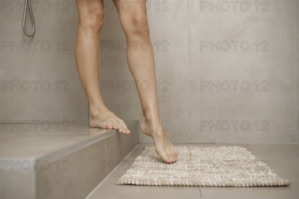 A person is carefully stepping out of a sleek shower onto a plush bath mat in a contemporary bathroom. The walls feature a neutral tone, enhancing the clean, minimalist aesthetic of the space