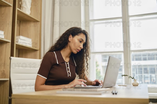In a cozy home office, a woman with long, curly hair focuses intently on her laptop while seated at a wooden desk