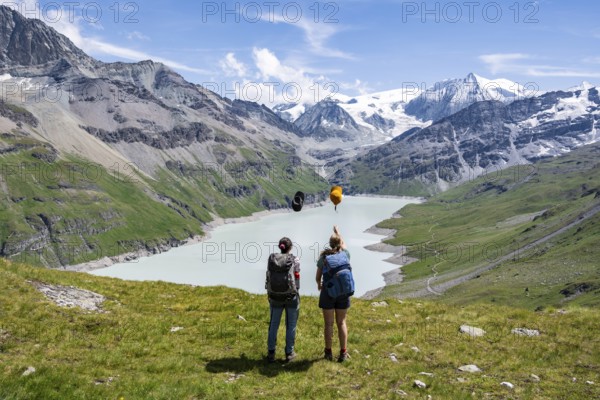 Two female friends throw their hats in the air, female mountaineers in front of a blue mountain lake, Lac des Dix reservoir, in the background mountain peak Mont Blanc de Cheilon with glacier, Hérménence, Valais Alps, Valais, Switzerland