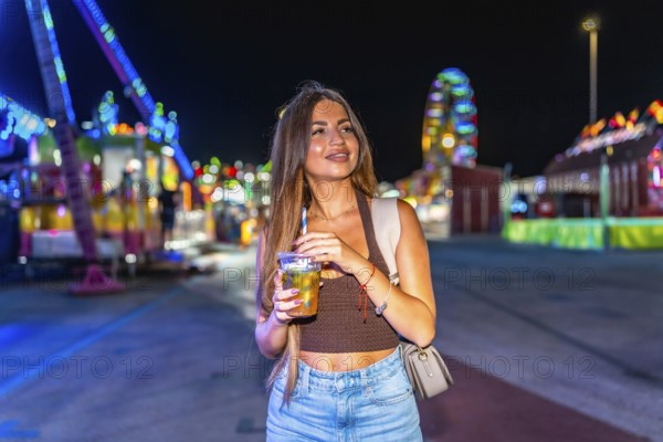 Young woman sips a beverage at a lively amusement park, illuminated by colorful lights, creating a joyful atmosphere