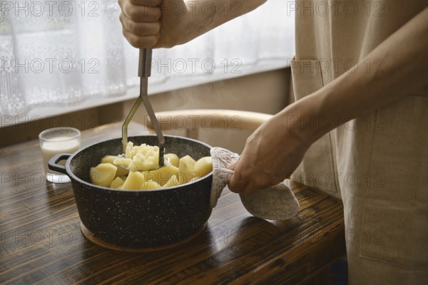 A person is mashing boiled potatoes in a pot. His hand grips a masher while holding a pot with another hand