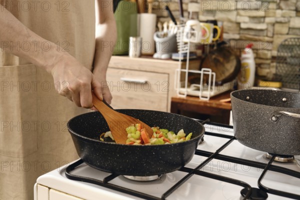 A person stirs a colorful mix of fresh vegetables in a frying pan on a stove in a cozy kitchen. Making vegetable base for ragout