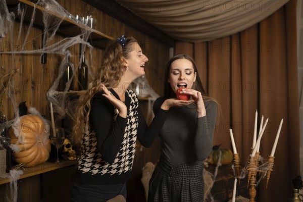 Two friends are playfully engaging in a Halloween celebration, surrounded by festive decorations. One friend holds a red apple while the other smiles and gestures in a cozy, spooky atmosphere