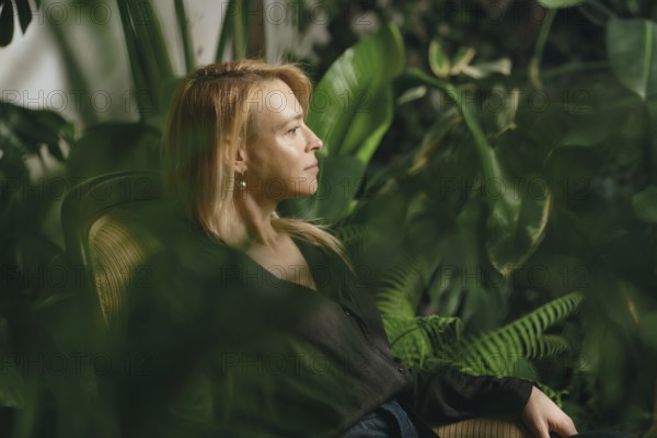 A woman sits quietly in a cozy chair within a vibrant indoor garden filled with lush plants and greenery. She appears thoughtful and relaxed, appreciating the tranquility of her surroundings
