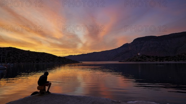 Angler sitting on the lakeshore during a colourful sunset against a mountainous backdrop, Holy Week, Bay of Panormitis, Panormitis Monastery, Symi, Dodecanese, Greek Islands, Greece